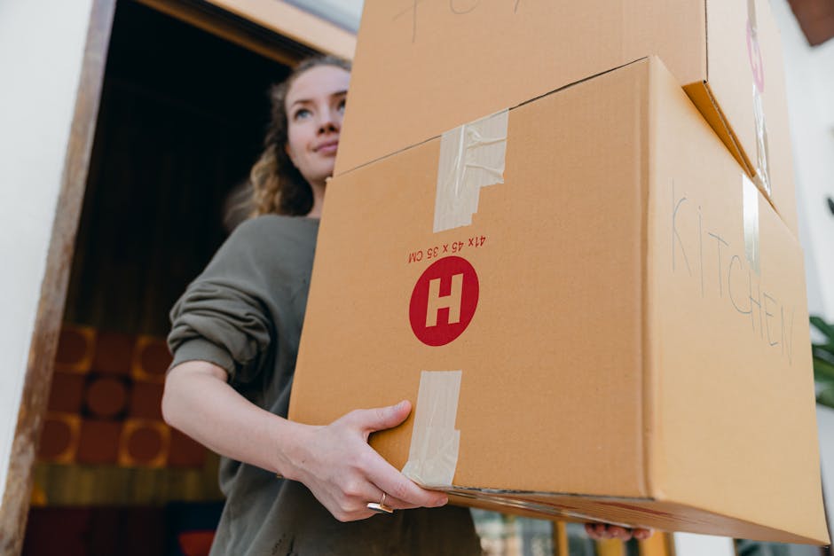 A woman with wavy brown hair, wearing a grey long-sleeved top, is inside a home near a doorway, carefully holding a medium-sized cardboard box with packaging tape and a red letter 'H' symbol, indicating it contains fragile or important items. The box has handwritten text on it, including the word 'KITCHEN', and appears to be part of a home relocation or moving process. Behind her, visible through the doorway, are multiple packed boxes and the interior of the house. The scene is well-lit with natural light, suggesting daytime, and the woman is engaged in lifting or positioning the box, which is likely prepared for transportation on a furniture or home relocation service, such as that offered by Man and Van Highbury. The environment reflects a typical move-in or move-out scenario involving packing materials, such as cardboard boxes secured with tape, readying for removal, with the focus on transporting household items safely during a house removal or relocation.