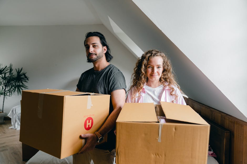 A man and a woman stand indoors on a wooden floor, each holding a large cardboard box with packing tape visible on top. The man has dark hair, a beard, and is wearing a casual grey t-shirt, standing slightly in front of the woman, who has curly blonde hair and is dressed in a pink and white striped shirt. They are positioned near a plain white wall with an angled ceiling, indicating an attic or loft space. In the background, a small potted plant is visible on the floor, and natural light illuminates the room, suggesting daytime. The scene appears to capture the process of home relocation, with the individuals preparing to load furniture or packed items, consistent with house removals and furniture transport services provided by [COMPANY_NAME], such as Man and Van Highbury, in the context of packing and moving. The image emphasizes the careful handling of packed belongings during a home move, reflecting professional packing and loading procedures.