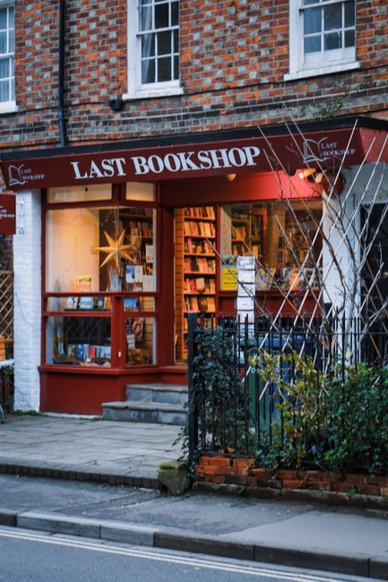 The exterior of a small bookshop named 'Last Bookshop' with a red sign and large display window showing shelves of books inside, illuminated by warm indoor lighting. The shop is set in a brick building with white-framed windows above. In front, there is a paved sidewalk leading to the entrance, which has a set of steps, and a black metal railing runs alongside a small garden area with shrubs and sparse foliage. The image captures a typical street scene during daylight, with no visible activity or people present. The house removals and furniture transport context is subtly implied through the storefront's position on the street, suggesting potential relocation activities nearby, as represented by the focus on shopfronts and urban environment.