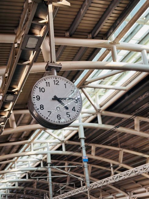A round analog clock mounted on a metal support fixture hangs from the ceiling of a busy train station or transit hub, with the time showing approximately 2:14. The station features a high, arched ceiling composed of white steel beams and transparent panels that allow natural light to illuminate the space. Visible structural elements include overhead electrical wiring and support trusses, indicating a modern, well-structured environment. The clock is positioned centrally in the image, with its white face, black numerals, and two black hands clearly distinguishable, providing an essential time reference for passengers and staff. In the background, components of the station’s infrastructure, such as additional steel beams and possibly signage, are visible but out of focus. This setting reflects a typical scene in a transportation hub where punctuality and efficient movement are priorities, aligning with the context of moving and logistics services offered by Man and Van Highbury, particularly related to house removals and furniture transport during home relocation processes.