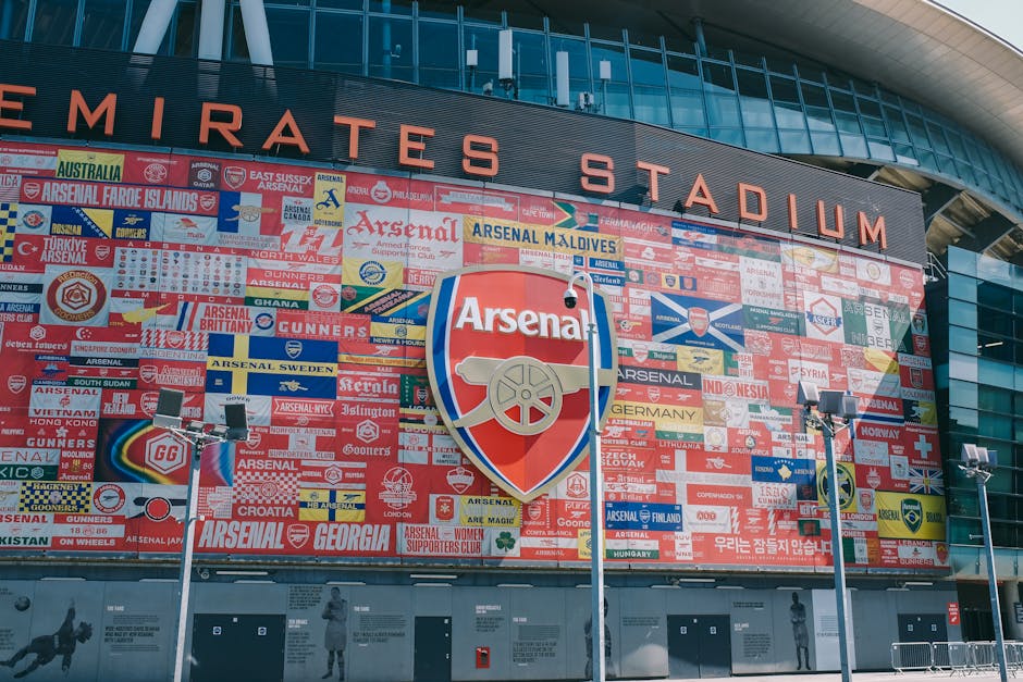 The exterior of the Emirates Stadium in Highbury features a large display of colorful banners and flags representing various Arsenal supporters' groups and countries, arranged on a dark panelled facad
