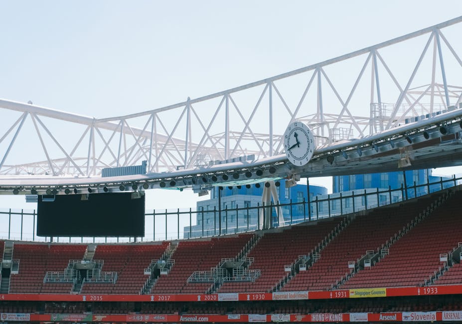 Inside a stadium, a section of red seating is visible with several cardboard boxes and plastic-wrapped furniture pieces arranged in a pile near a doorway. Some items are being lifted or carried by a m