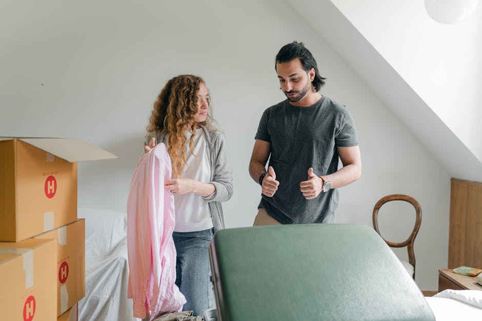 A scene inside a home showing two individuals preparing for a house move or furniture transport. On the left, a woman with curly red hair is holding a pink and white striped fabric cover, possibly a protective cover for a piece of furniture or a box. She is dressed in casual clothing, wearing a light gray cardigan over a white top. On the right, a man with dark hair tied back, wearing a dark gray t-shirt and beige trousers, is giving a thumbs-up gesture, indicating readiness or approval. In front of them, there is a large green padded moving blanket draped over a piece of furniture to protect it during the relocation process. To the left, several cardboard boxes with red markings are stacked on a white sofa, some still open and packed with packaging materials. The room has a simple, neutral-colored interior with natural light coming through a window, and a wooden chair is visible in the background. This scene illustrates the packing and moving preparations involved in a residential home relocation, with [COMPANY_NAME] providing furniture transport and removal services as part of house removals in the N5, Highbury area.