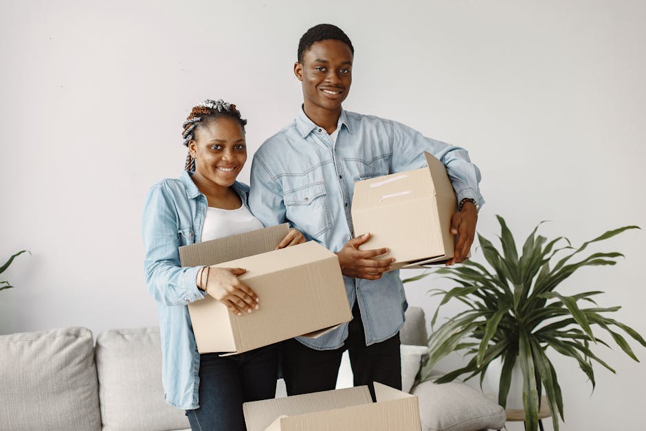 A young couple stands indoors, each holding cardboard moving boxes filled with household items, as part of a home relocation process. The woman on the left has a smile, short curly hair styled with accessories, and is wearing a light denim jacket over a white top. The man on the right is also smiling, with short black hair, and is dressed in a light blue button-up shirt. They are positioned in front of a plain white wall with natural lighting illuminating the scene. Behind them, there is a grey sofa with a beige cushion, and a large green houseplant with broad leaves situated to the right. In the foreground, a partially opened cardboard box on a table or surface is visible, suggesting they are in the process of packing or unpacking. This image exemplifies furniture transport, packing and moving activities, associated with professional removals services offered by Man and Van Highbury, supporting home relocation and logistics in the N5, Highbury area.