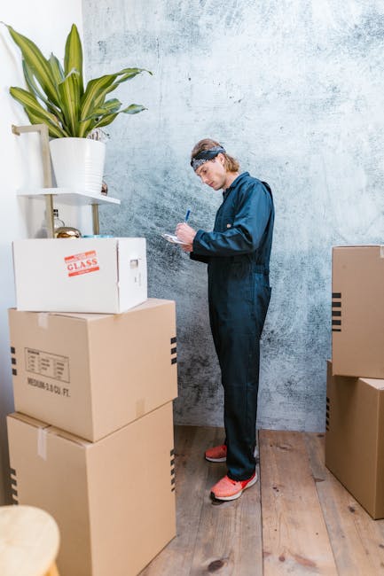 A man wearing a blue protective jumpsuit, headband, and orange safety shoes is standing indoors against a textured light blue wall, appearing to assess or record details on a clipboard. He is surrounded by several cardboard boxes of varying sizes, some sealed with packing tape and labels, including one marked 'GLASS,' indicating fragile items. The boxes are stacked on a wooden floor, with additional boxes placed nearby, suggesting a packing or moving process. A large potted plant with long green leaves sits on a white shelf to the left, contrasting with the utilitarian scene. The environment appears to be a residential interior prepared for a house relocation or furniture transport, and the man and van service provider, Man and Van Highbury, is likely involved in planning or managing the move. This scene illustrates an organized packing or loading stage as part of a professional removals service within a home or flat in the N5, Highbury area.
