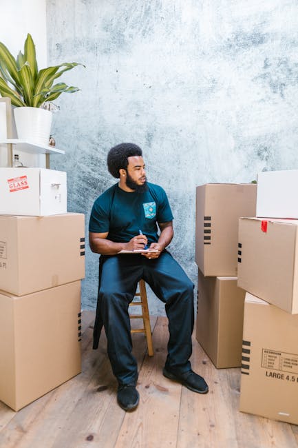 A man wearing a blue protective jumpsuit, headband, and orange safety shoes is standing indoors against a textured light blue wall, appearing to assess or record details on a clipboard. He is surrounded by several cardboard boxes of varying sizes, some sealed with packing tape and labels, including one marked 'GLASS,' indicating fragile items. The boxes are stacked on a wooden floor, with additional boxes placed nearby, suggesting a packing or moving process. A large potted plant with long green leaves sits on a white shelf to the left, contrasting with the utilitarian scene. The environment appears to be a residential interior prepared for a house relocation or furniture transport, and the man and van service provider, Man and Van Highbury, is likely involved in planning or managing the move. This scene illustrates an organized packing or loading stage as part of a professional removals service within a home or flat in the N5, Highbury area.