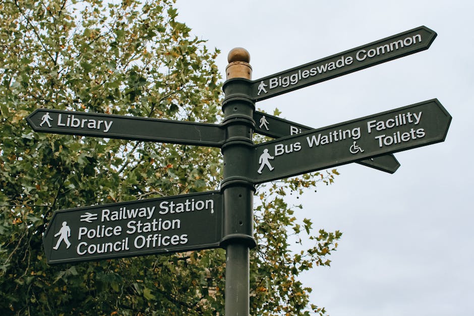 A black multi-directional street signpost situated outdoors, attached to a central pole with four horizontal arms, each pointing in different directions. The top arm indicates 'Biggleswade Common', the second arm points to 'Library', the third points to 'Bus Waiting Facility' and 'Toilets', and the bottom arm directs to 'Railway Station', 'Police Station', and 'Council Offices'. The signs feature white text and icons, with pedestrian symbols and accessibility symbols. In the background, there are green leafy trees and a partly cloudy sky, suggesting the scene is in a public area or community space. The image may be used in contexts related to local navigation, planning house relocations near public amenities, or movement logistics in Highbury, supporting topics on efficient house removals and transportation coordination, as covered in the 'Islington Council permit guide for Highbury removals and skips' page by Man and Van Highbury.
