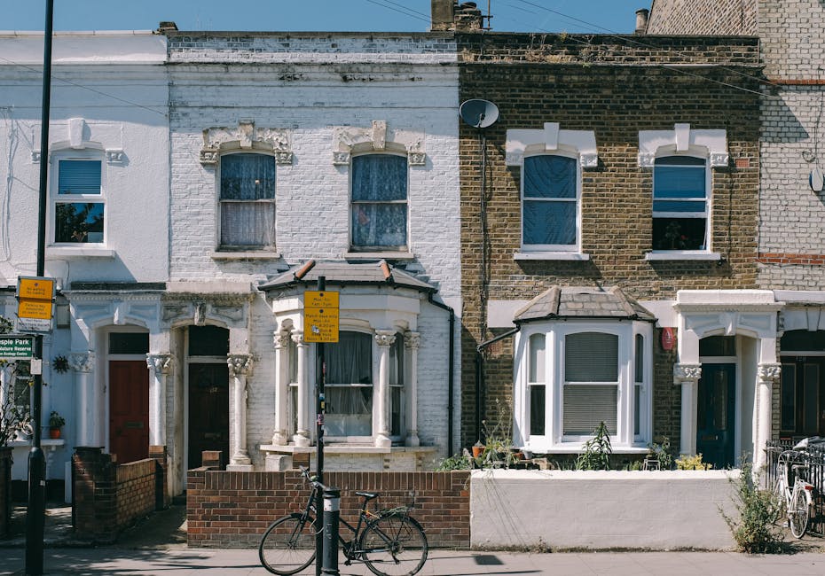 A row of three terraced residential houses with Victorian-style architectural features, including bay windows and decorative columns at the entrance. The leftmost house has a white painted facade with a red front door and a small front garden enclosed by a low brick wall. In front of the houses, a black bicycle is parked against one of the lamp posts, with two yellow parking restriction signs attached. The middle house has a white facade with two upper windows and a bay window on the ground floor, containing various potted plants and garden items. The rightmost house has a brick facade with large front windows and a small garden area. The scene is illuminated by daylight, with the pavement in front of the houses supporting typical urban street features such as plant pots and bicycles. The overall setting suggests a quiet residential area suitable for house removals and home relocation activities, with relevant signage and a typical London street environment. Man and Van Highbury occasionally operates in this area for local removals and transport services, emphasizing professional packing, loading, and furniture transport processes.