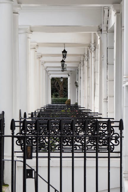 A long, narrow covered porch with white-painted wooden columns and beams supporting a ceiling, illuminated by hanging black lantern-style light fixtures. The porch is enclosed by a decorative black wr