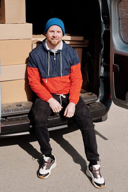 A man wearing a blue beanie, a navy and red waterproof jacket, black trousers, and white trainers with black and red details sits on the edge of an open van door, smiling at the camera. Inside the van