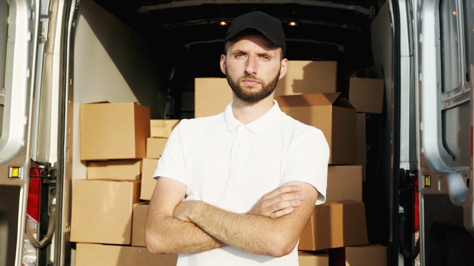 A man wearing a blue beanie, a navy and red waterproof jacket, black trousers, and white trainers with black and red details sits on the edge of an open van door, smiling at the camera. Inside the van