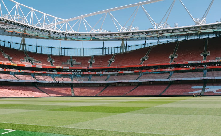 Inside a large sports stadium, the playing field is visible with well-maintained grass divided into sections by mowing patterns. Surrounding the field are multiple tiers of red and pink seating sections, which are mostly empty. Above the seating area, a modern white steel roof structure with a network of beams and supports stretches across the top, providing partial shade and shelter for spectators. The sky overhead is clear with a few clouds, and bright sunlight illuminates the entire scene. This setting indicates a venue suitable for hosting football matches or other sports events, and the image captures the interior environment where the playing field and seating areas are prepared for an event, such as a home football fixture or a large sports gathering. As part of a house removal or moving service, such an area might be used for illustrative purposes related to relocating or transporting items for an event venue or sports team.