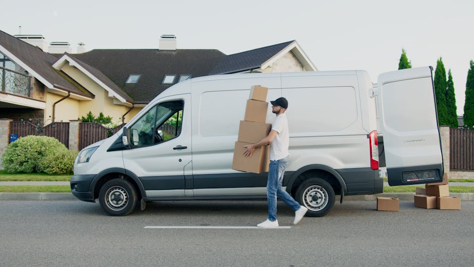 A man wearing a white t-shirt, blue jeans, and a black cap is walking alongside a white commercial van with its side door open on a residential street. He is carrying four cardboard boxes stacked in his arms, and several additional boxes are placed on the pavement near the van. Inside the van, visible through the open side door, are more packed cardboard boxes and some larger items wrapped in protective materials, likely prepared for removal or home relocation. The van is parked in front of a modern, two-storey house with a dark roof, light-colored walls, and a wooden fence with greenery, including trees and bushes, in the background. The scene appears to be part of the loading process involved in packing and moving furniture or household items, aligned with professional removals services provided by Man and Van Highbury.